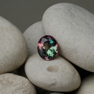 A multicolored, oval-cut alexandrite gemstone with shades of pink, green, and blue sits on smooth, white pebbles, creating an elegant and tranquil scene.