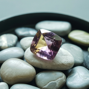 A faceted, bi-color ametrine gemstone with lavender and yellow hues sits atop smooth, gray pebbles. The scene is calm and elegant, highlighting the gem's sparkle.