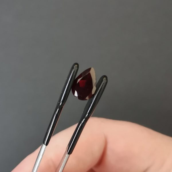 The deep red garnet teardrop gem stone being held in tweezers against a black background