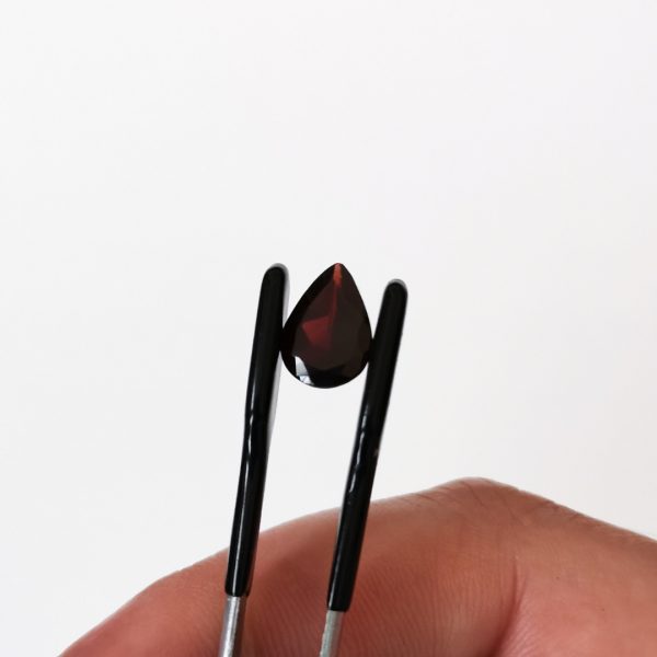 The deep red garnet teardrop gemstone being held in tweezers against a white background