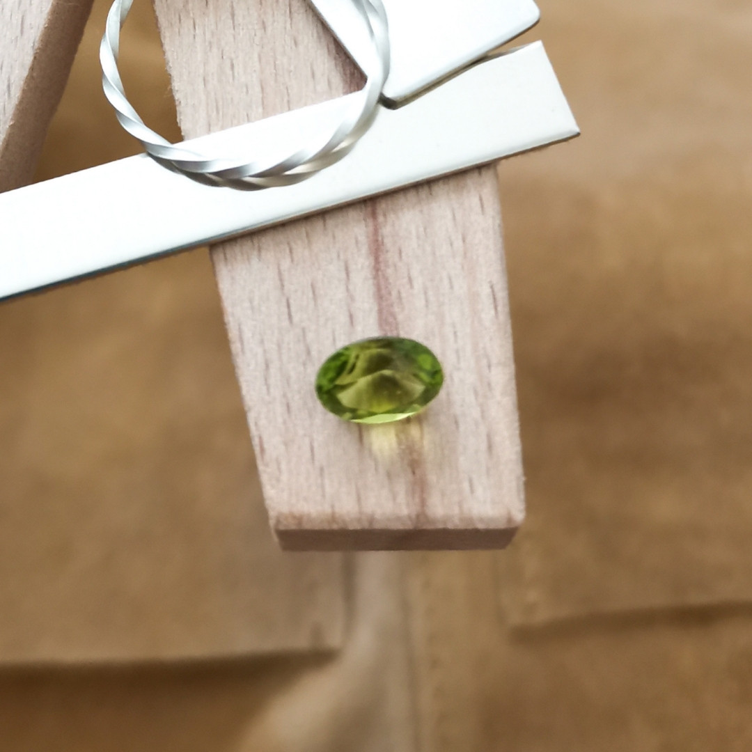 A loose green peridot gemstone in a faceted oval cut resting on a jewellers bench peg by some silver