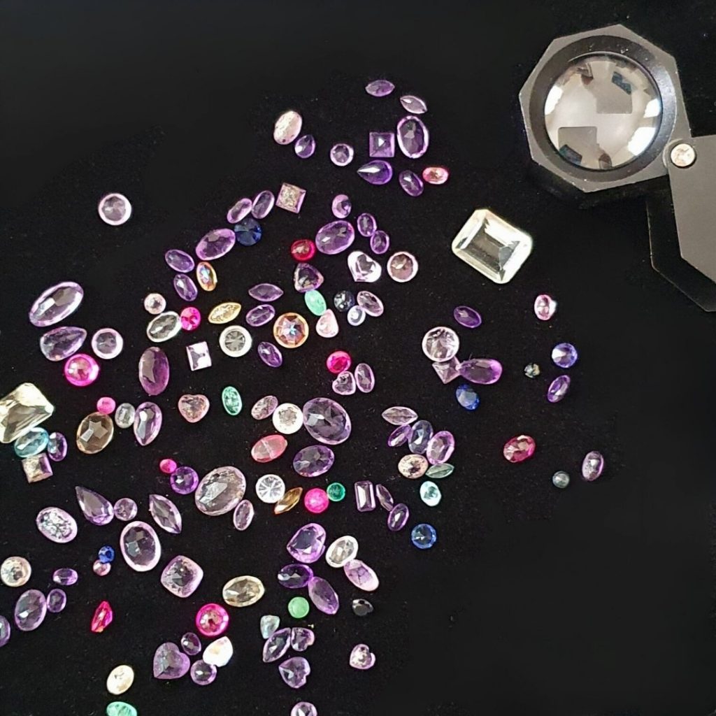 A photograph of coloured reclaimed and recycled gemstones scattered across a black background with a jewellers loupe laying next to them