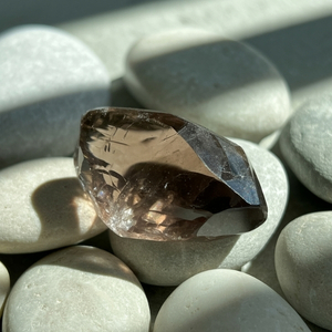 A faceted, transparent brown smoky quartz gemstone rests on smooth white pebbles, with sunlight casting soft shadows, creating a serene and tranquil feel.