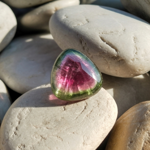 A triangular, polished pink and green watermelon tourmaline gemstone sits on smooth white stones, with sunlight casting a soft shadow, creating a calm, natural ambiance.