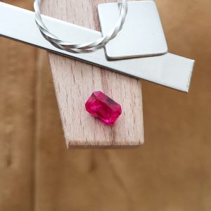 The pinkish red lab grown ruby rectangle displayed on a jewellers bench peg with some sterling silver sheet and wire.