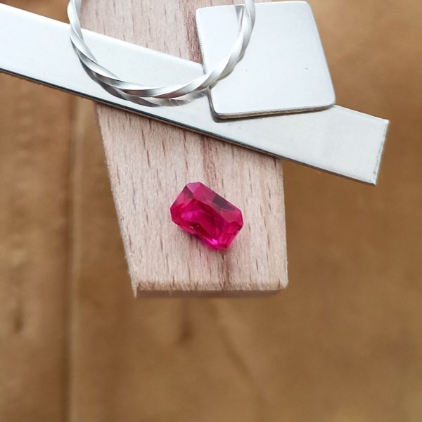 The pinkish red lab grown ruby rectangle displayed on a jewellers bench peg with some sterling silver sheet and wire.