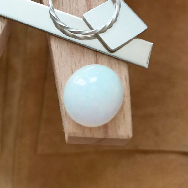 The white opal oval cabochon displayed on a jewellers bench peg with some sterling silver sheet and wire.