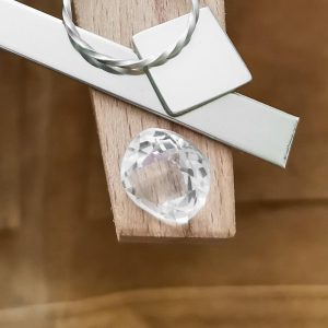 The cushion cut square clear quartz displayed on a jewellers bench peg with some sterling silver sheet and wire.