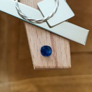 The cushion cut square blue sapphire displayed on a jewellers bench peg with some sterling silver sheet and wire.
