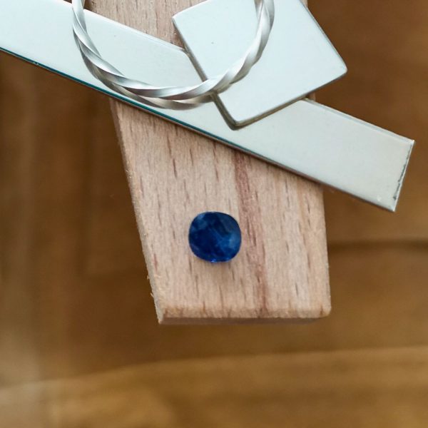 The cushion cut square blue sapphire displayed on a jewellers bench peg with some sterling silver sheet and wire.
