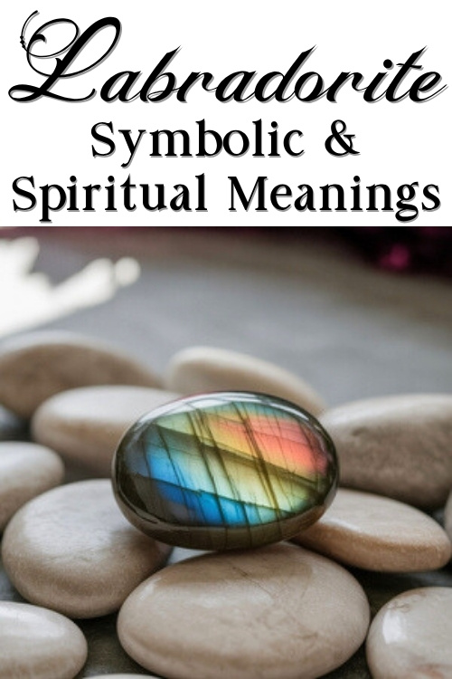 A polished labradorite stone rests on smooth beige pebbles, displaying a vibrant blue, yellow, and red sheen. Text above reads "Labradorite: Symbolic & Spiritual Meanings."