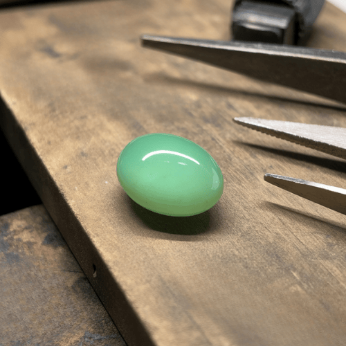 A smooth, oval green Chrysoprase gemstone sits on a wooden workbench, surrounded by precision tools.