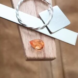 The teardrop cut orange hessonite garnet displayed on a jewellers bench peg with some sterling silver sheet and wire.