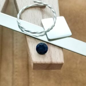 The round cut blue lab grown sapphire displayed on a jewellers bench peg with some sterling silver sheet and wire.