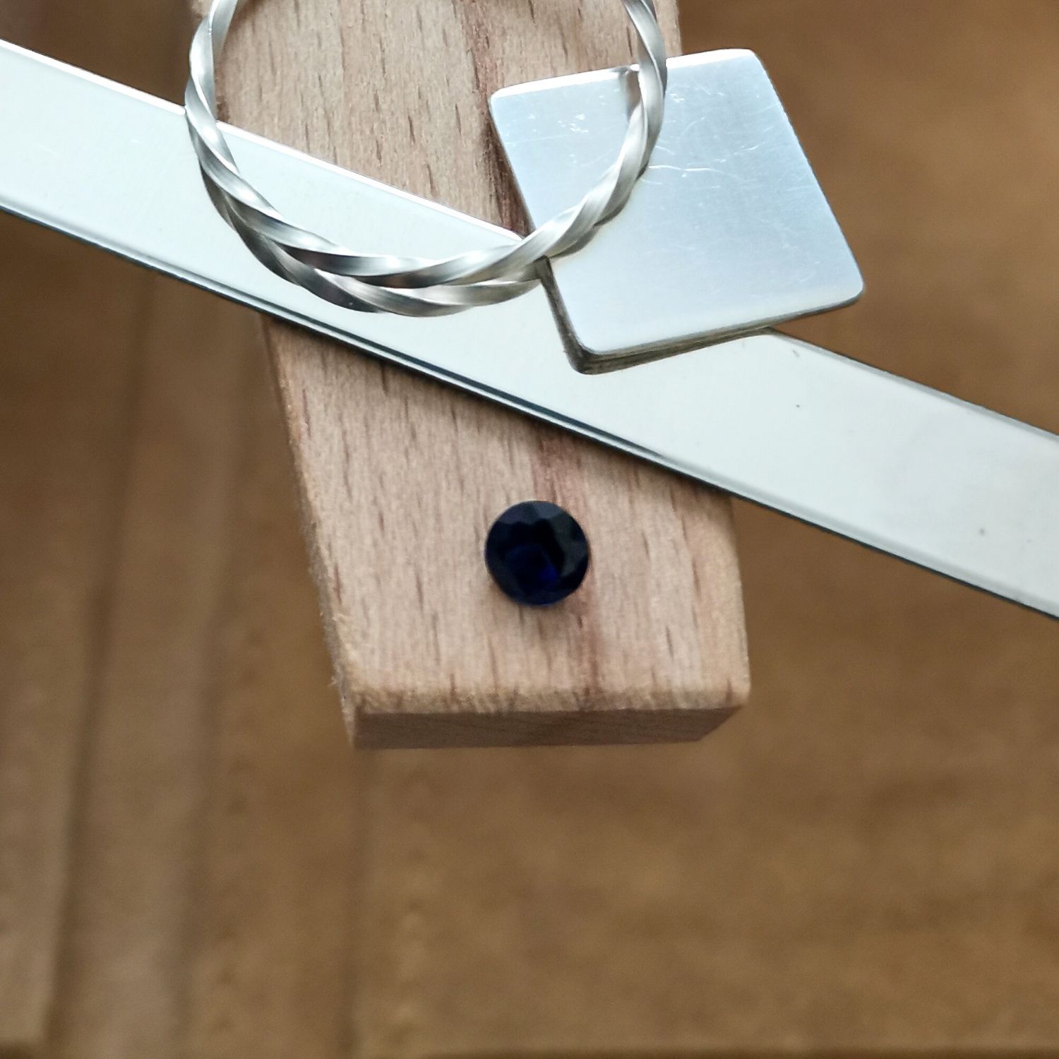 The small round cut blue lab grown sapphire displayed on a jewellers bench peg with some sterling silver sheet and wire.