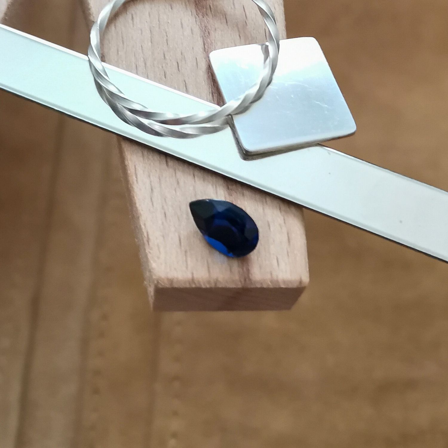The teardrop cut blue lab grown sapphire displayed on a jewellers bench peg with some sterling silver sheet and wire.