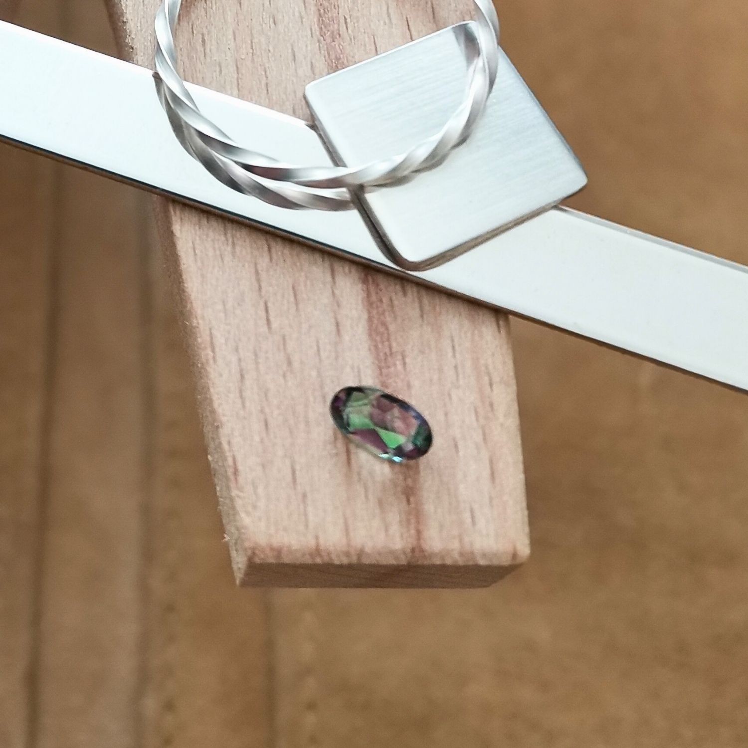 The facet cut oval mystic topaz displayed on a jewellers bench peg with some sterling silver sheet and wire.