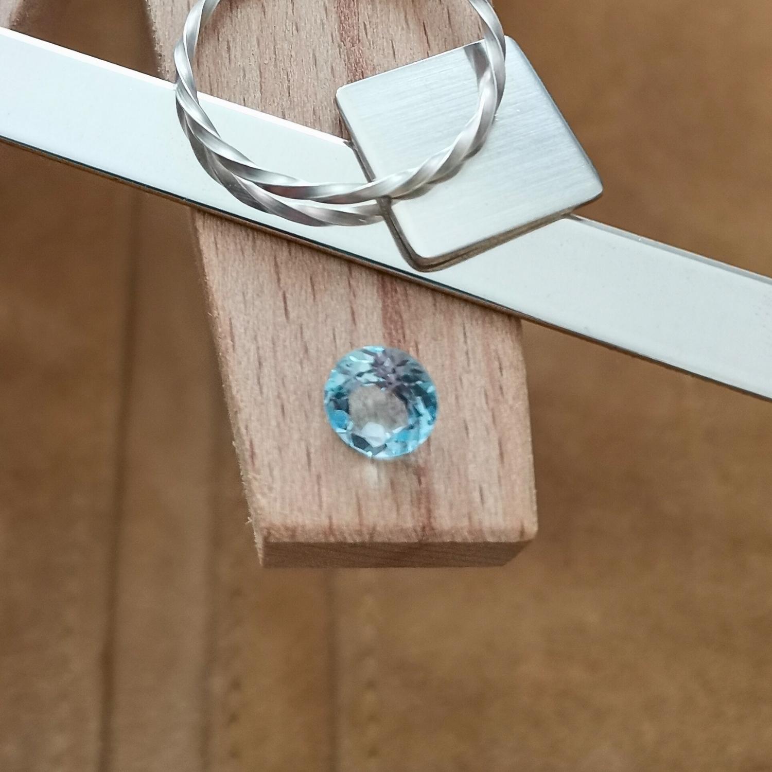 The blue topaz that's been facet cut into a circle is displayed on a jewellers bench peg with some sterling silver sheet and wire.