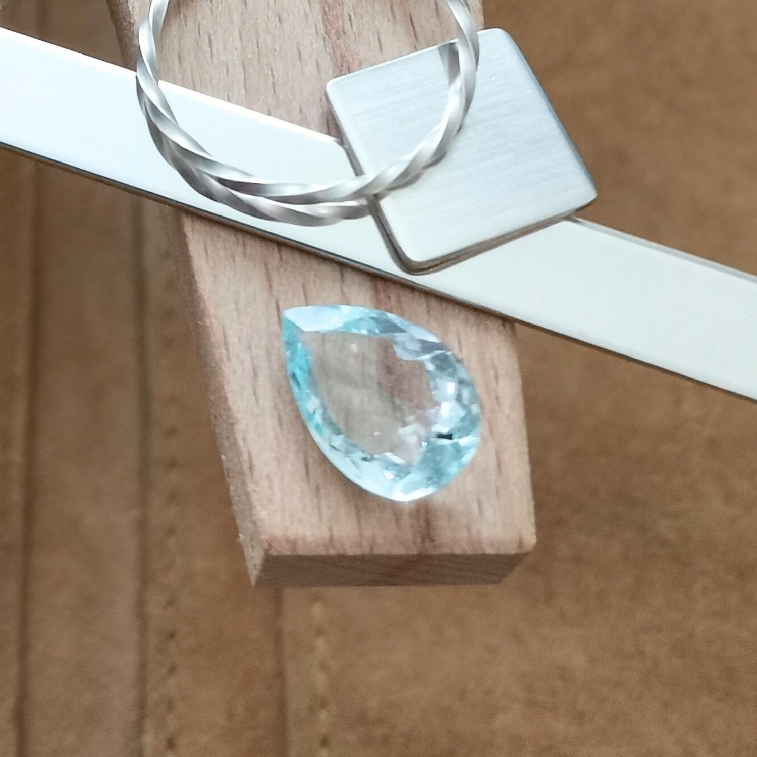 The facet cut aquamarine teardrop displayed on a jewellers bench peg with some sterling silver sheet and wire.