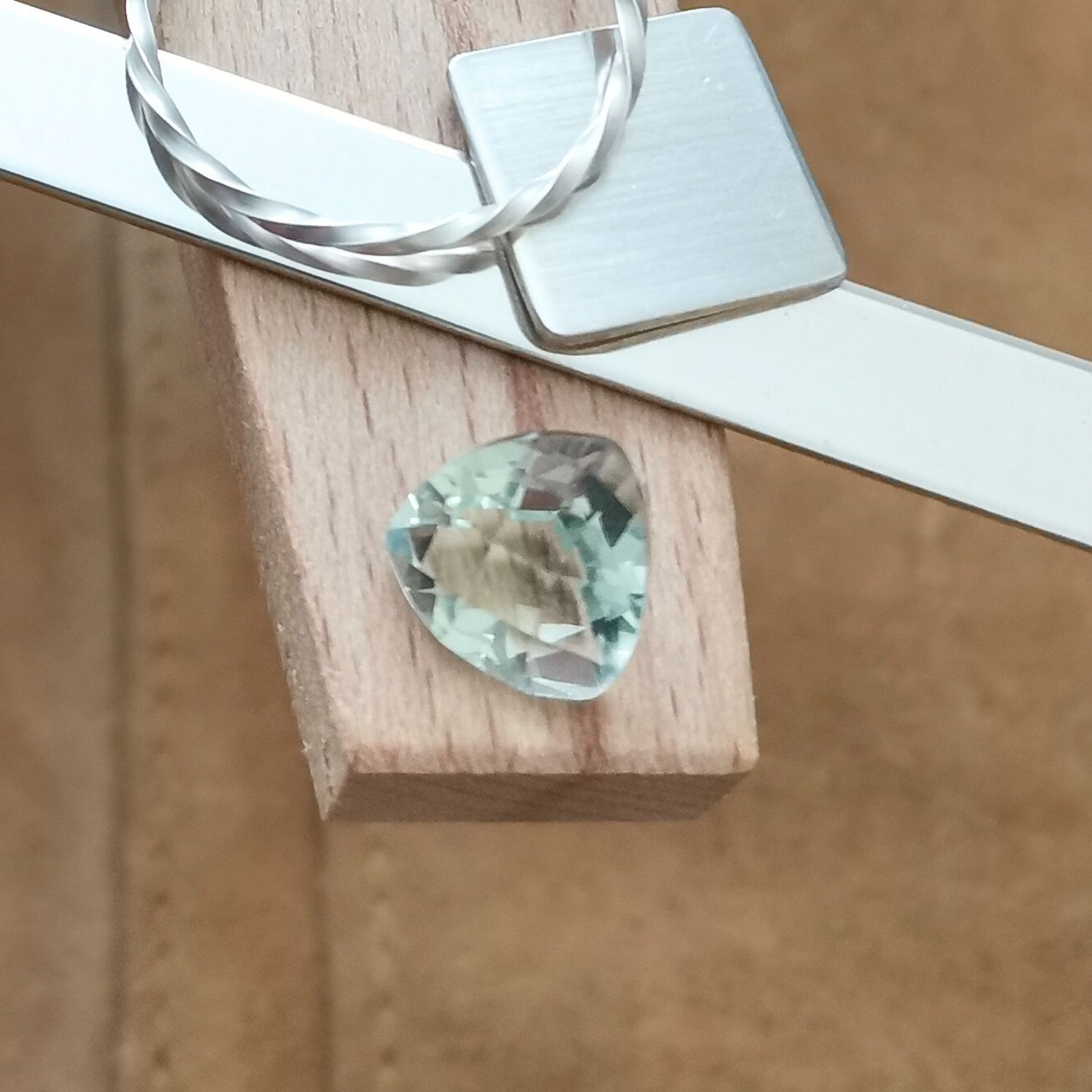 The fancy facet cut pale green prasiolite triangle displayed on a jewellers bench peg with some sterling silver sheet and wire.
