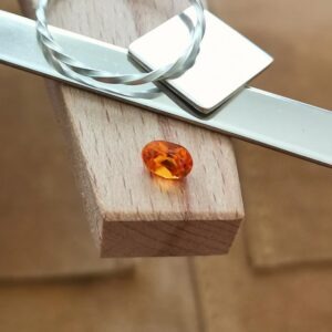 The bright orange facet cut lab grown sapphire oval displayed on a jewellers bench peg with some sterling silver sheet and wire.