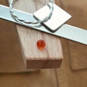 The small orange faceted round garnet displayed on a jewellers bench peg with some sterling silver sheet and wire.