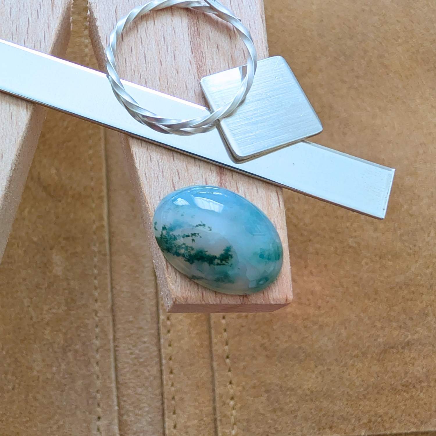 The loose oval moss agate cabochon is being displayed on a wooden jewellers bench peg with some sterling silver sheet and wire.