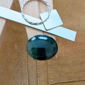 The loose oval moss agate cabochon is being displayed on a wooden jewellers bench peg with some sterling silver sheet and wire.