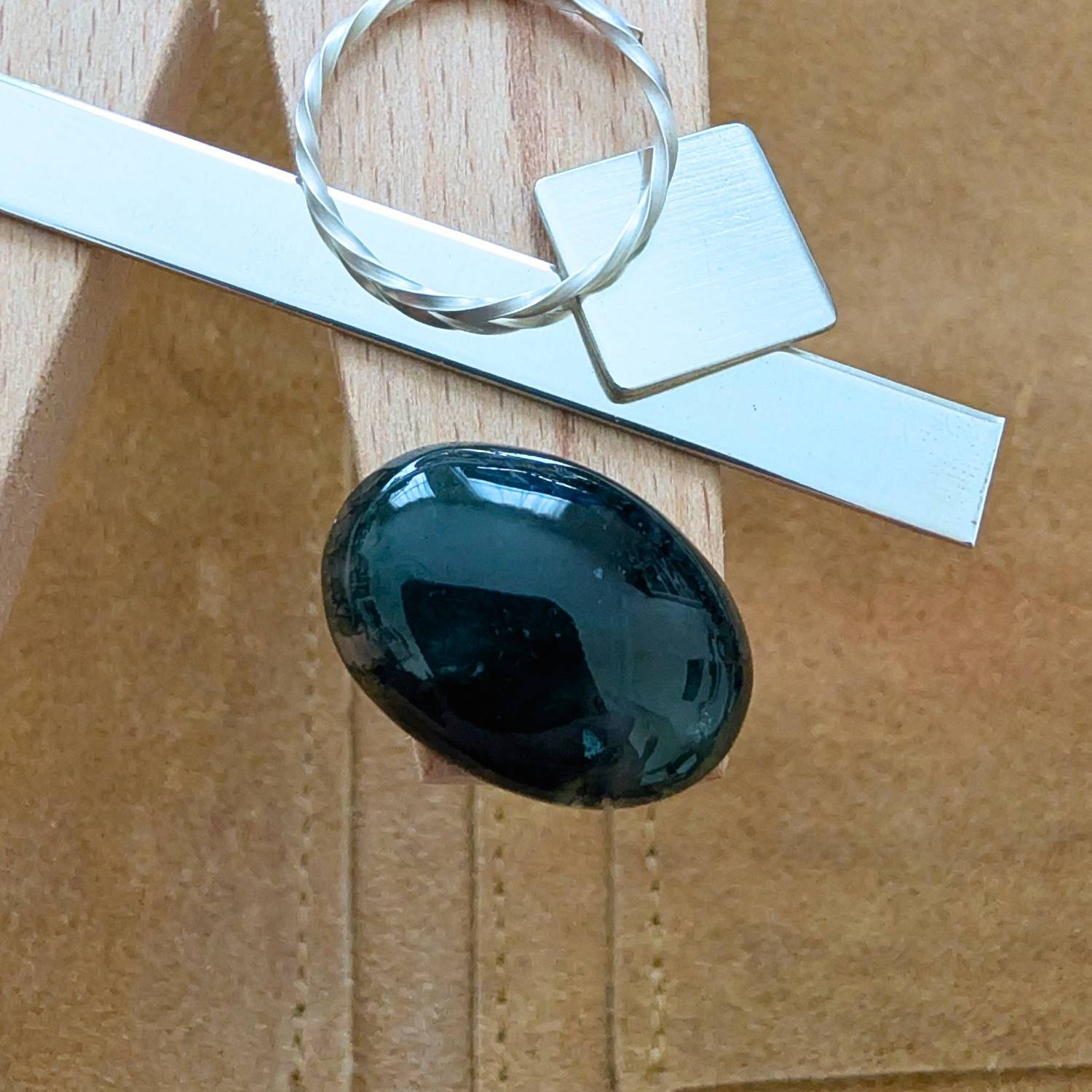 The loose oval moss agate cabochon is being displayed on a wooden jewellers bench peg with some sterling silver sheet and wire.