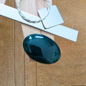 The loose oval moss agate cabochon is being displayed on a wooden jewellers bench peg with some sterling silver sheet and wire.