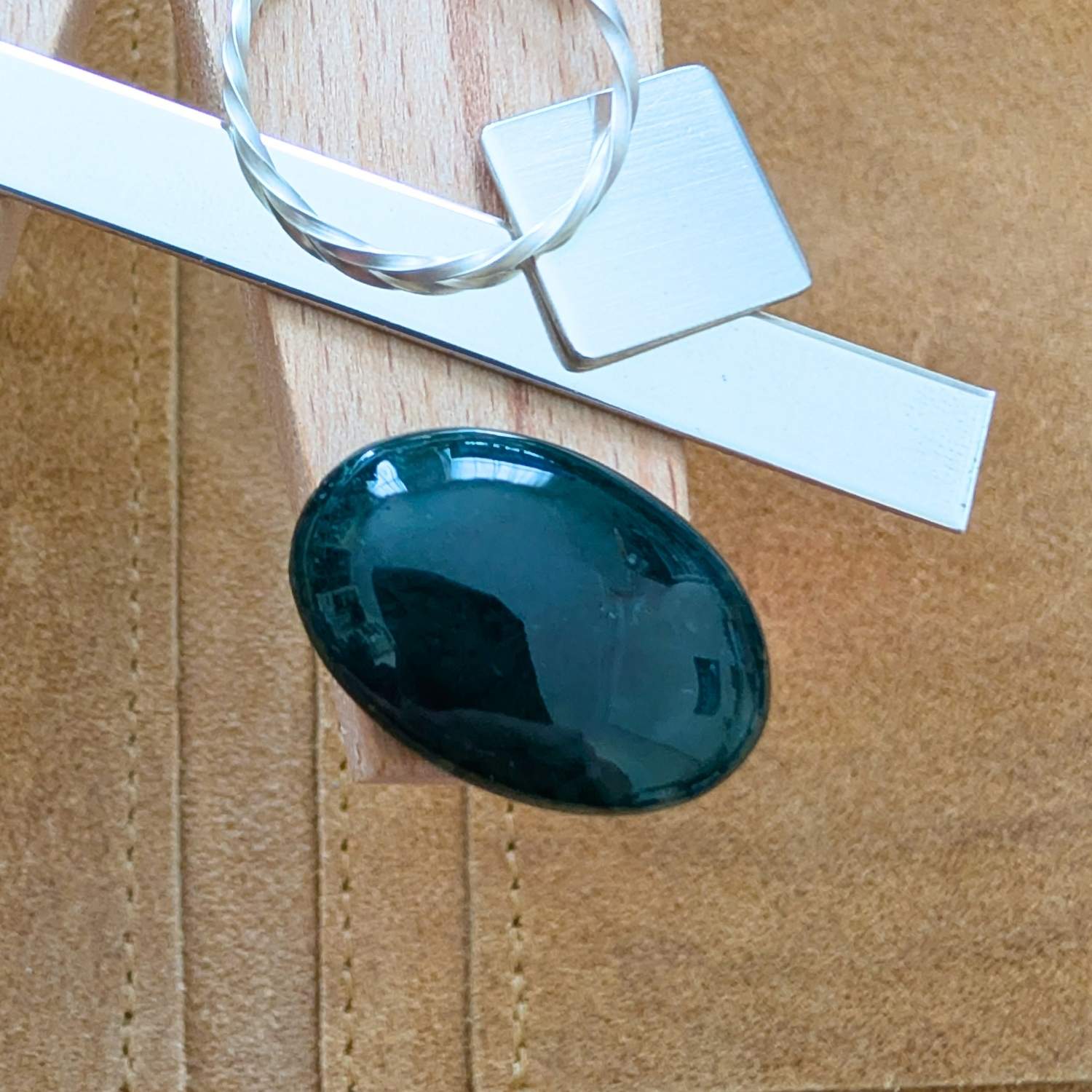 The loose oval moss agate cabochon is being displayed on a wooden jewellers bench peg with some sterling silver sheet and wire.