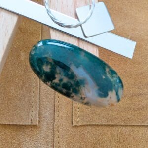 The loose oval moss agate cabochon is being displayed on a wooden jewellers bench peg with some sterling silver sheet and wire.