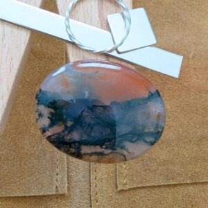 The loose oval moss agate cabochon is being displayed on a wooden jewellers bench peg with some sterling silver sheet and wire.