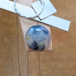 The loose round dendritic agate cabochon is being displayed on a wooden jewellers bench peg with some sterling silver sheet and wire.