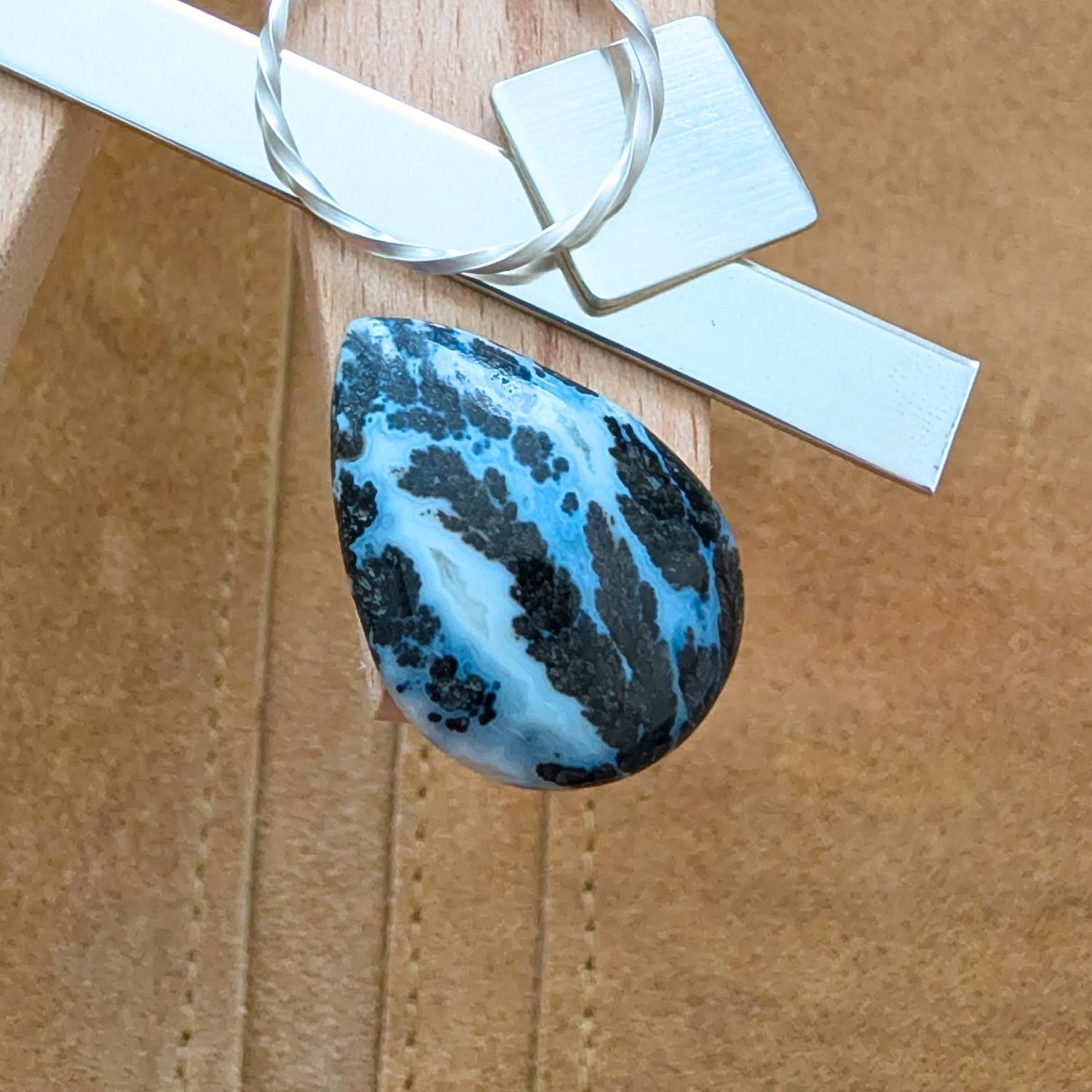 The loose teardrop dendritic agate cabochon is being displayed on a wooden jewellers bench peg with some sterling silver sheet and wire.