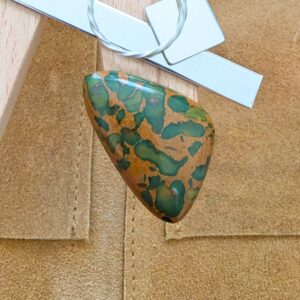 The loose freeform fruit jasper cabochon is being displayed on a wooden jewellers bench peg with some sterling silver sheet and wire.