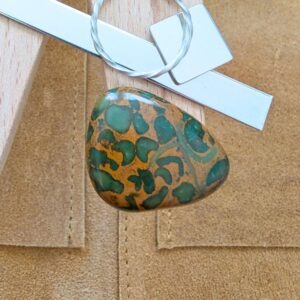 The loose freeform fruit jasper cabochon is being displayed on a wooden jewellers bench peg with some sterling silver sheet and wire.