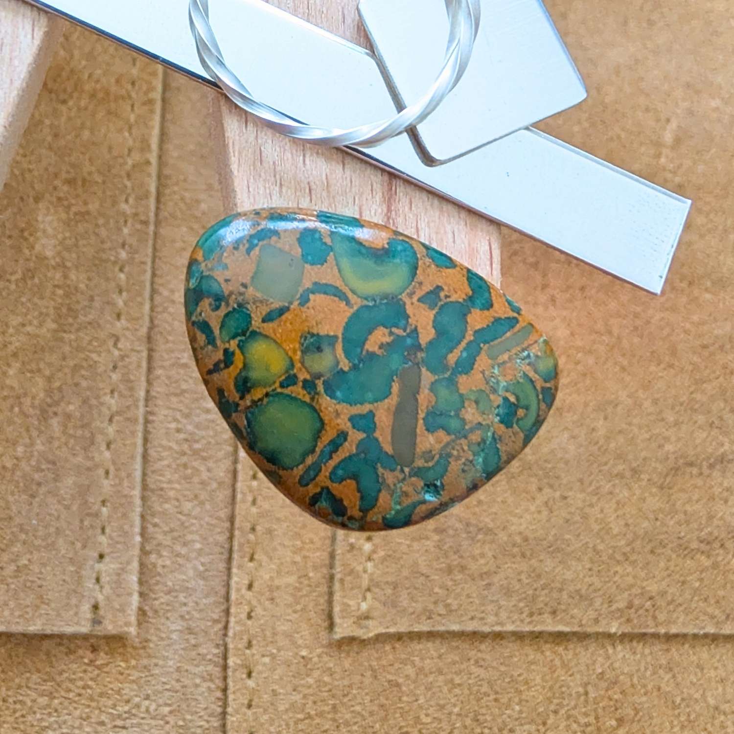 The loose freeform fruit jasper cabochon is being displayed on a wooden jewellers bench peg with some sterling silver sheet and wire.