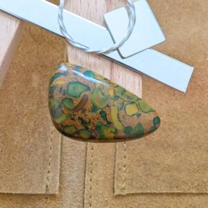The loose freeform fruit jasper cabochon is being displayed on a wooden jewellers bench peg with some sterling silver sheet and wire.