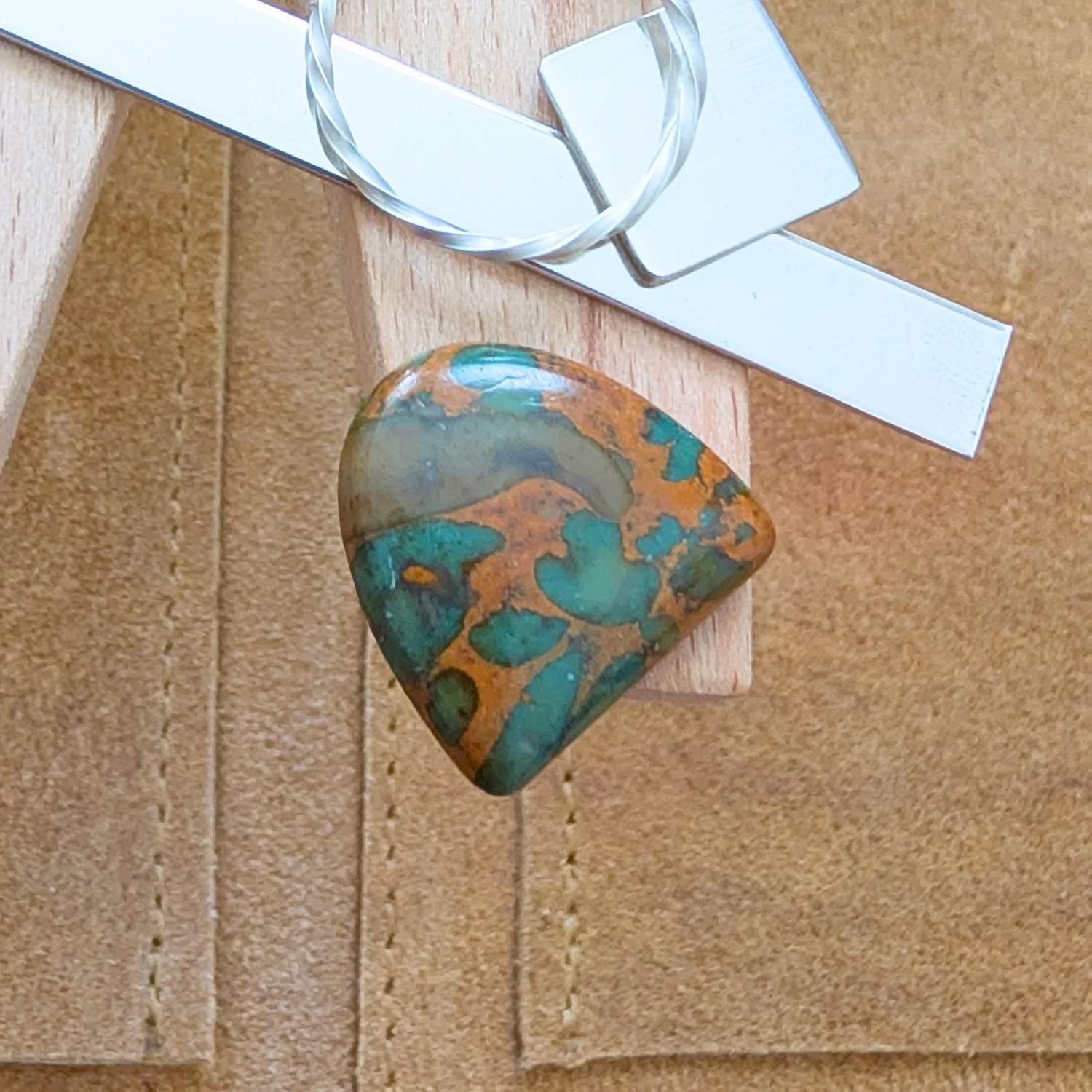 The loose freeform fruit jasper cabochon is being displayed on a wooden jewellers bench peg with some sterling silver sheet and wire.