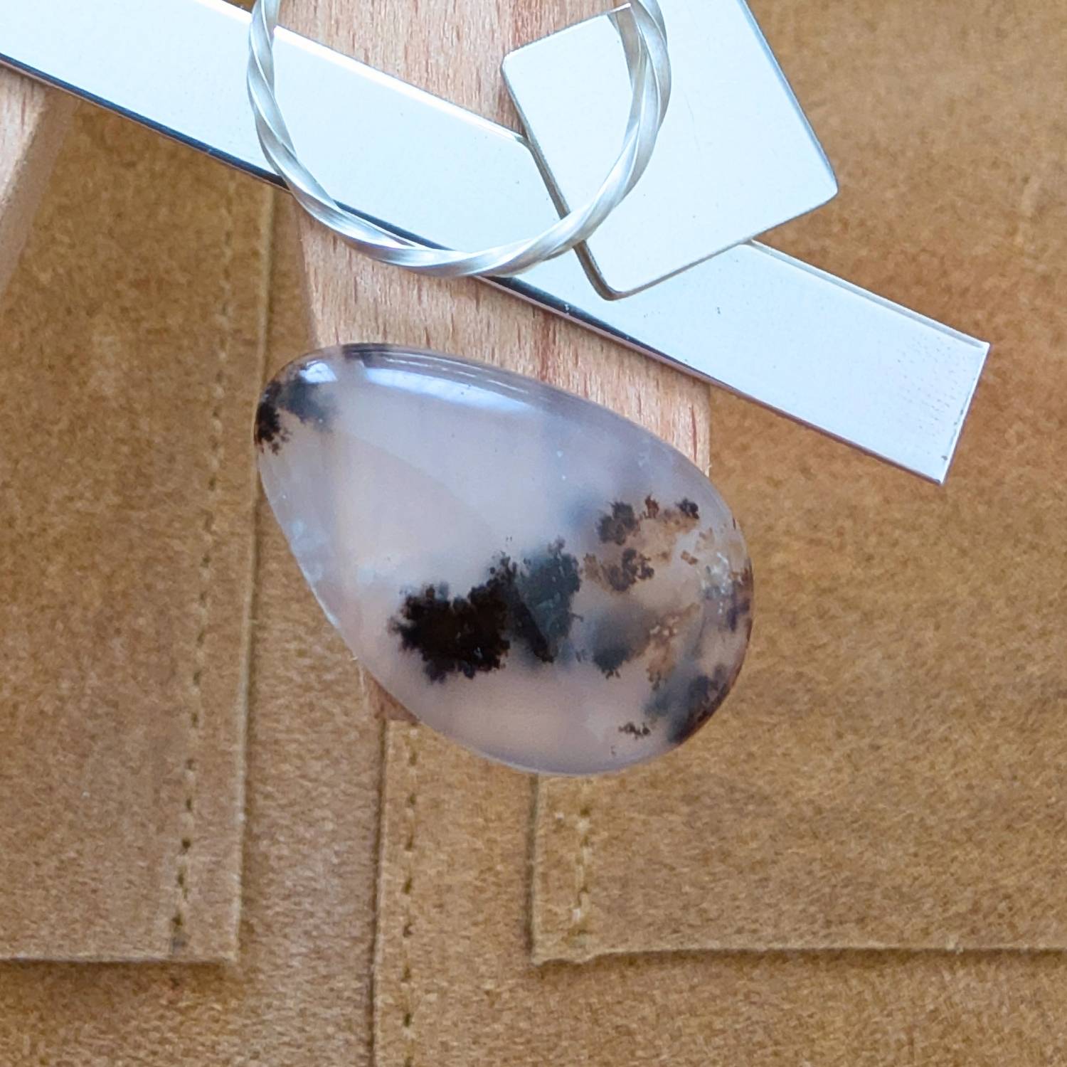 The loose freeform dendritic opal cabochon is being displayed on a wooden jewellers bench peg with some sterling silver sheet and wire.