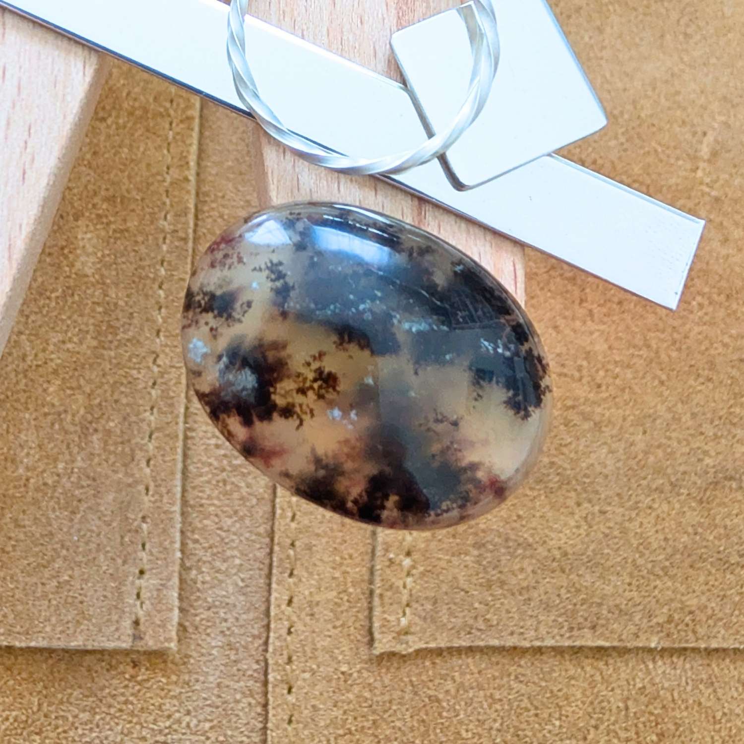 The loose dendritic opal cabochon is being displayed on a wooden jewellers bench peg with some sterling silver sheet and wire.