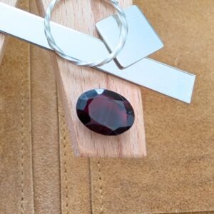 The loose faceted red garnet oval is being displayed on a wooden jewellers bench peg with some sterling silver sheet and wire.