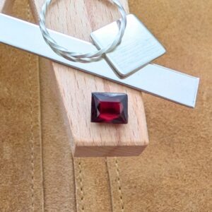 The loose faceted red garnet rectangle is being displayed on a wooden jewellers bench peg with some sterling silver sheet and wire.