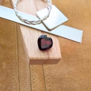 The loose faceted red garnet heart is being displayed on a wooden jewellers bench peg with some sterling silver sheet and wire.