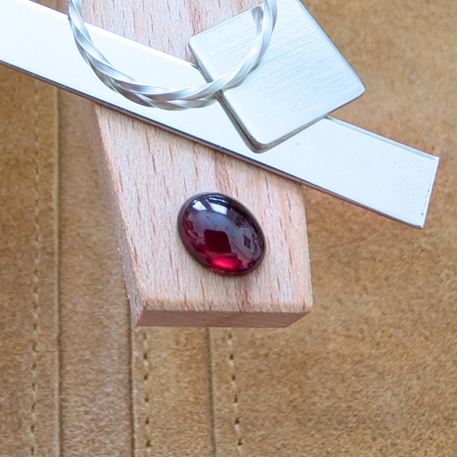 The loose red garnet oval cabochon is being displayed on a wooden jewellers bench peg with some sterling silver sheet and wire.