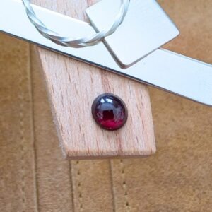 The loose red garnet round cabochon is being displayed on a wooden jewellers bench peg with some sterling silver sheet and wire.