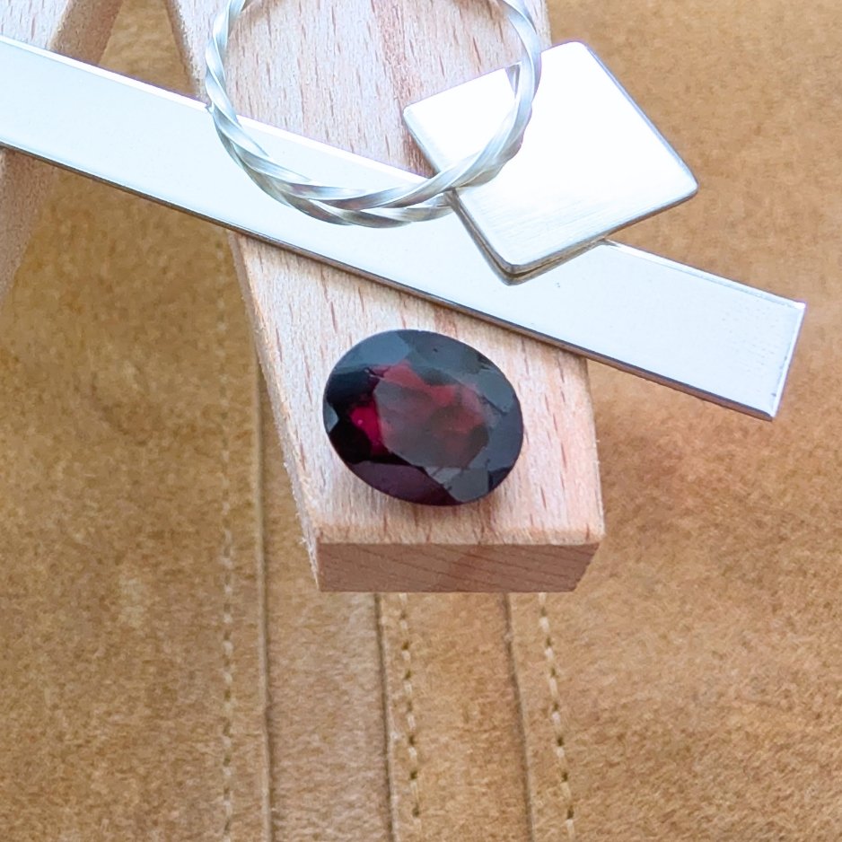 The loose faceted red garnet oval is being displayed on a wooden jewellers bench peg with some sterling silver sheet and wire.
