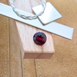 The loose faceted red garnet round is being displayed on a wooden jewellers bench peg with some sterling silver sheet and wire.