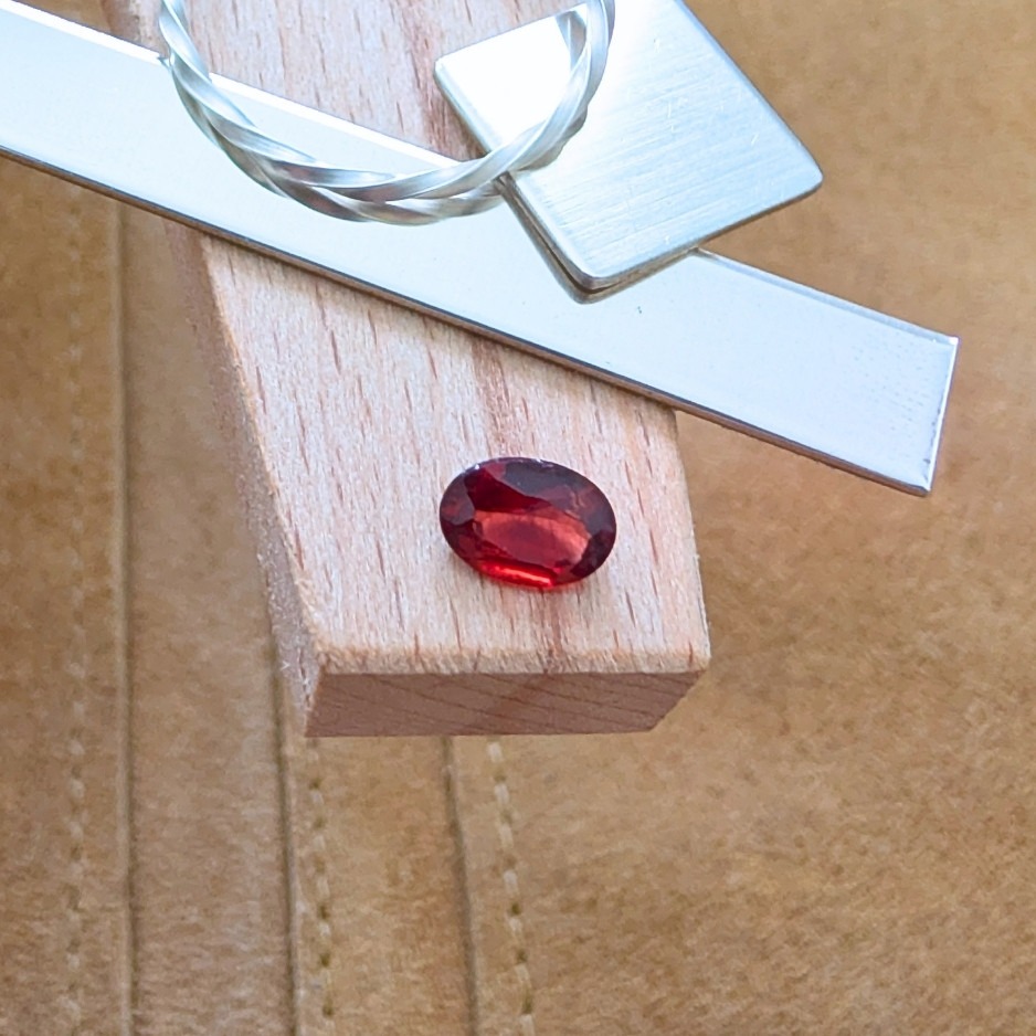 The loose faceted red garnet oval is being displayed on a wooden jewellers bench peg with some sterling silver sheet and wire.